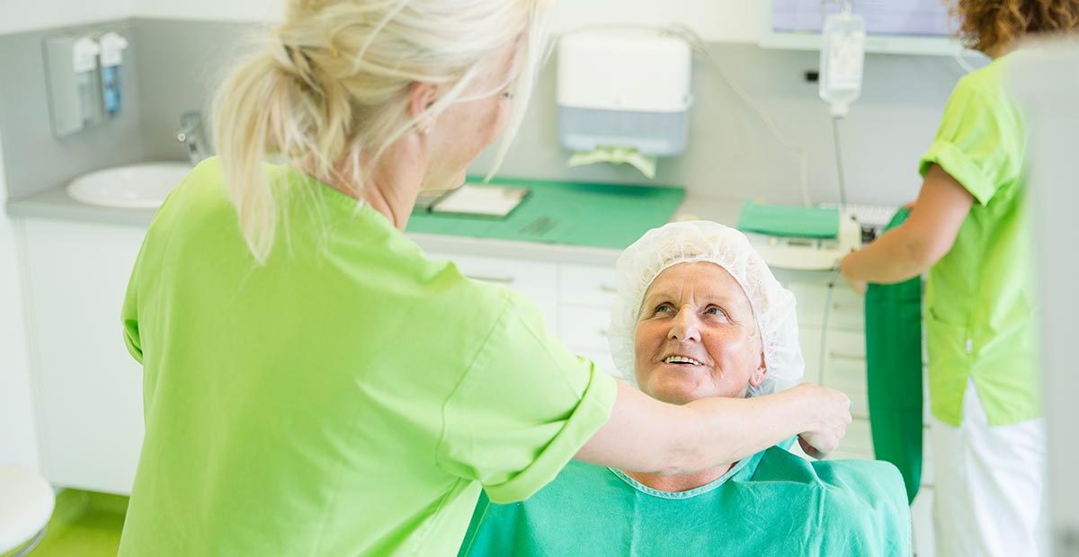 Eine Mitarbeiterin legt einer älteren Frau im Behandlungsstuhl einen OP-Umhang um, während sie angelächelt wird. Im Hintergrund bereitet eine andere Mitarbeiterin einen weiteren OP-Kittel vor.