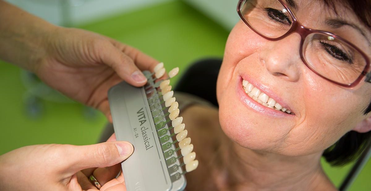 Zwei Frauenhände halten Beispiele für Zahnfarbe an die Zähne einer Frau, die lächelt