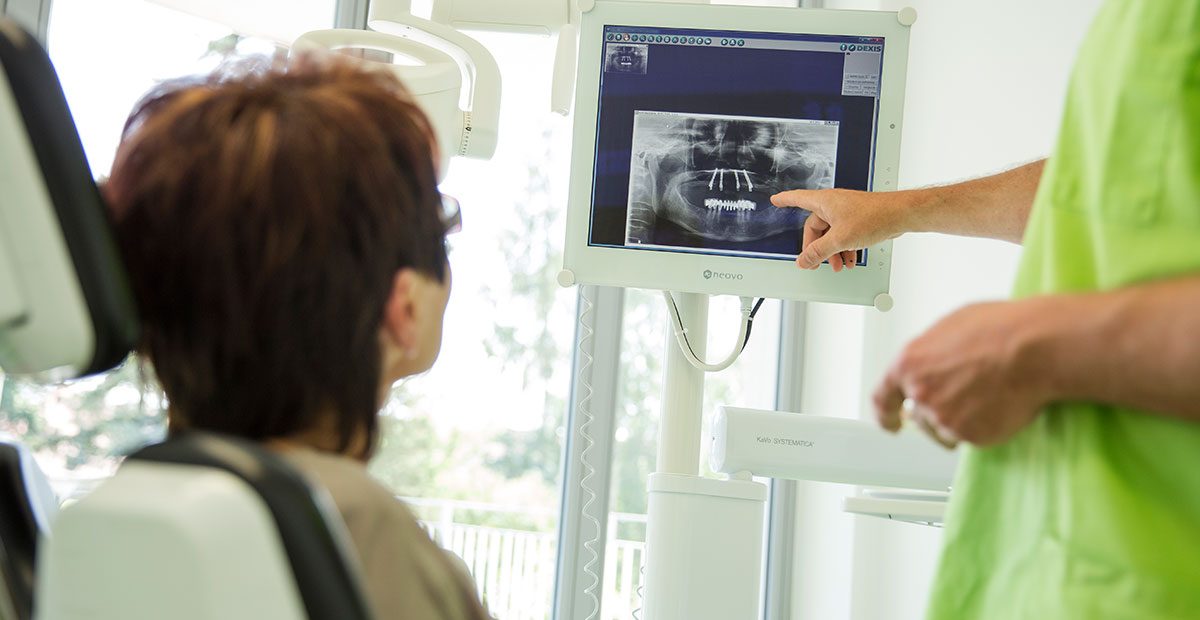 Männerhand zeigt auf eine Röntgenaufnahme und erklärt einer Patientin etwas, die im Zahnarztstuhl sitzt.