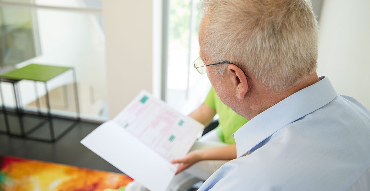 Älterer Patient ist von hinten zu sehen, der im Heil- und Kostenplan etwas nachliest, während eine Mitarbeiterin im Hintergrund den Heil- und Kostenplan hoch hält.