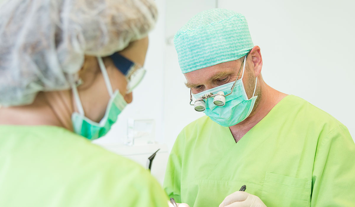 Zahnarzt Dr. Thomas Buchmann hat einen Mundschutz an und trägt eine OP-Haube sowie zwei Vergrößerungslinsen. Im Vordergrund ist eine Mitarbeiterin zu sehen, die ebenfalls Mundschutz trägt.
