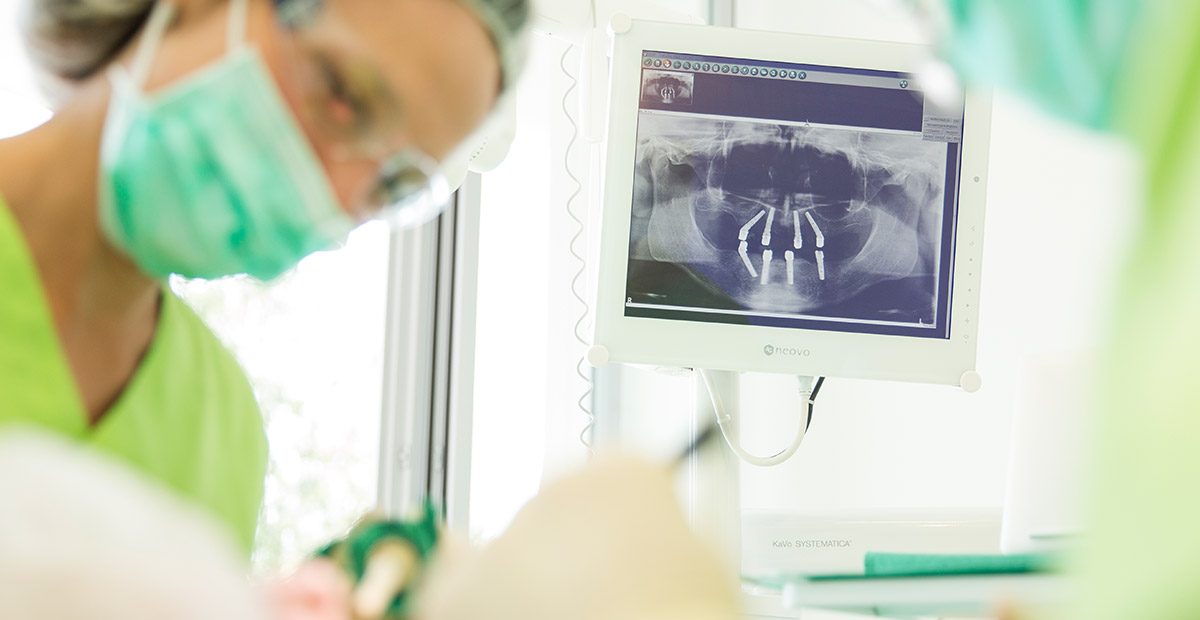 Mitarbeiterin mit Mundschutz beugt sich vornüber und hält Zahnarztinstrumente in der Hand, im Vordergrund ist eine andere Person mit Mundschutz zu sehen. Im Hintergrund sieht man ein Röntgenbild mit Zahnimplantaten.