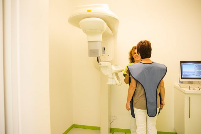 Patientin und Mitarbeiterin stehen sich gegenüber, links daneben ist ein Digitaler Volumentomograph zu sehen. Die Patientin trägt eine Bleischürze, die Mitarbeiterin lächelt sie an.
