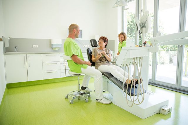 Zahnarzt Dr. Thomas Buchmann sitzt neben einer Frau im Behandlungsstuhl, die ihm etwas erklärt. Im Hintergrund sitzt Cornelia Renneberg und lächelt.