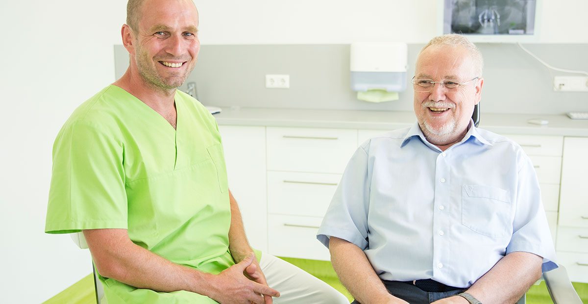Zahnarzt Dr. Thomas Buchmann sitzt auf einem Stuhl im Behandlungszimmer und lächelt, rechts daneben sitzt ein älterer Herr und lacht ebenfalls. Im Hintergrund sind Röntgenbilder zu sehen.