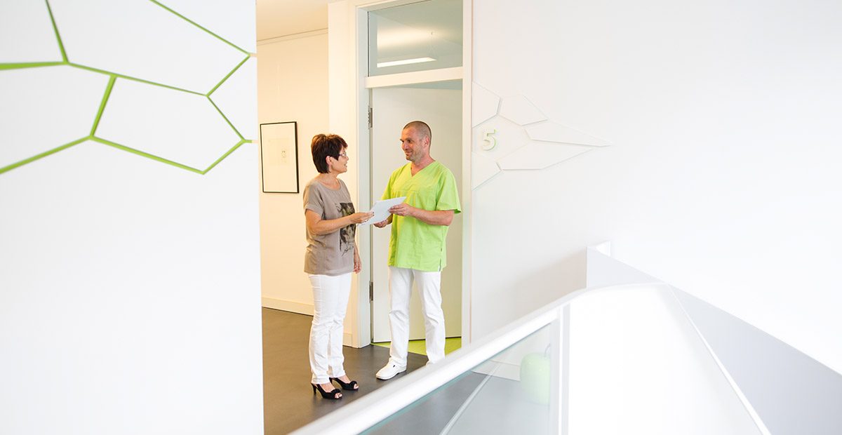 Dr. Thomas Buchmann steht mit einer Patientin vor einer Tür und gibt ihr einen Vorsorgeplan in die Hand. Beide schauen sich an und lächeln.