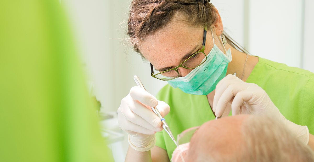 Zahnärztin Karin Kurze trägt einen Mundschutz und hält in beiden Händen Zahnarztbesteck, während Patient im Stuhl liegt, von dem nur der Kopf zu sehen ist.