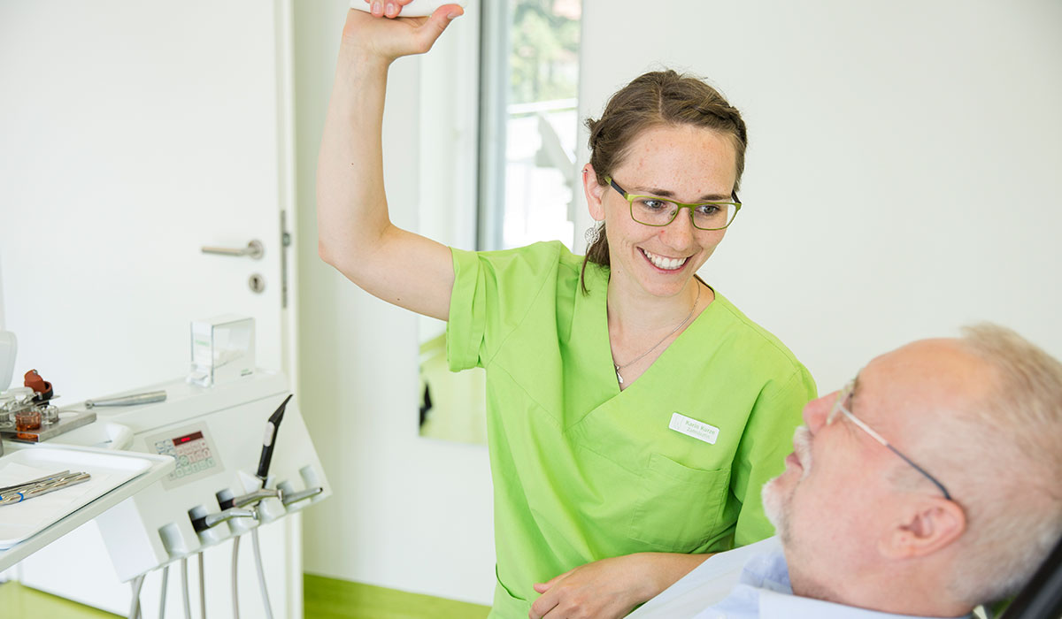 Zahnärztin Karin Kurze lächelt älteren Herrn im Zahnarztstuhl an, während sie das Behandlungslicht mit der rechten Hand ausrichtet. Der Patient liegt im Stuhl und lächelt sie an.