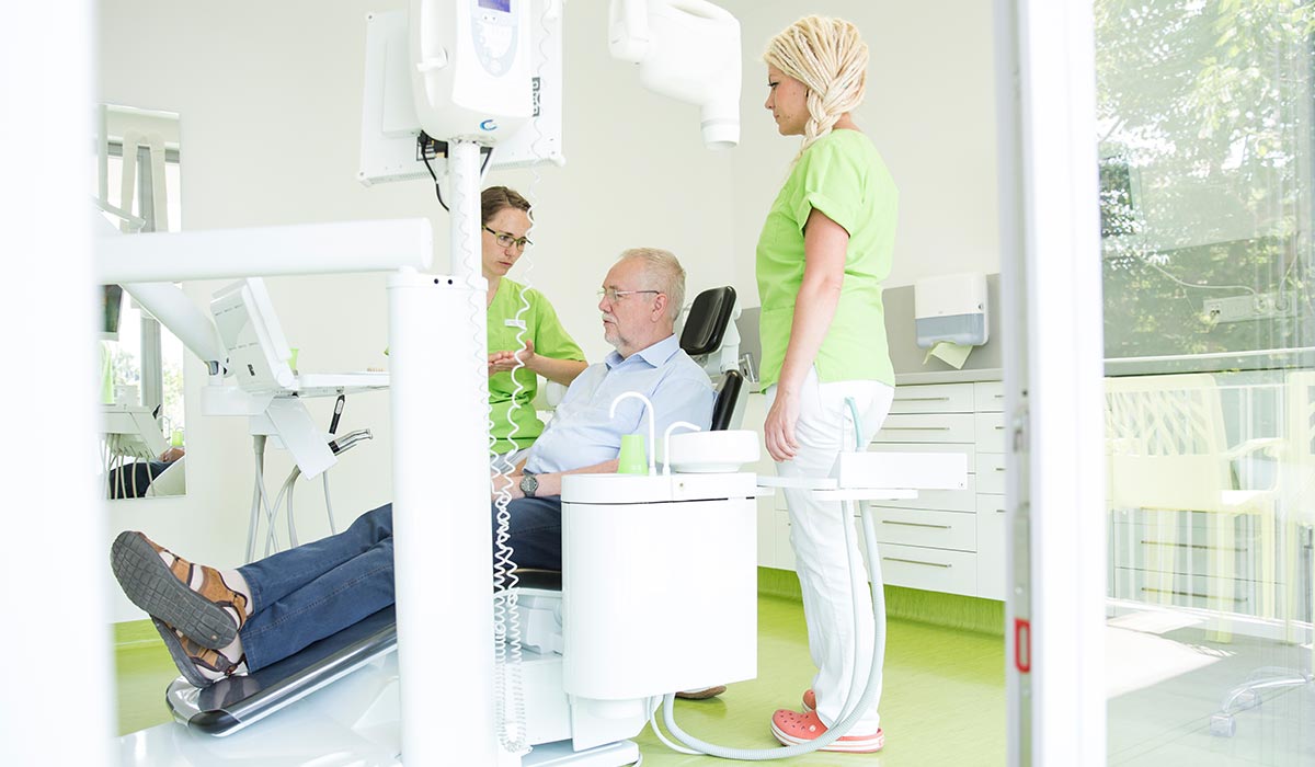 Zahnärztin Karin Kurze sitzt neben älterem Patienten im Stuhl und erklärt ihm etwas, während eine Mitarbeiterin daneben steht.